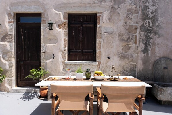 An outdoor dining area with a stone wall, two wooden doors, a table with chairs, and various food items. - The Fabrica Falasarna (Kissamos)