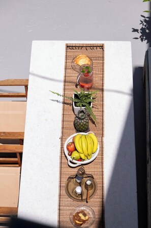 A table with a placemat, a glass pitcher, a plate of bananas, a plate of apples, a small potted plant, a decorative pineapple, a glass of water, and a cup with a spoon. - The Fabrica Falasarna (Kissamos)