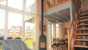 A spacious living room with a large window, a striped armchair, a fireplace, a dining area, and a wooden staircase. - Tolles Ferienhaus fur Familien (Rechlin)