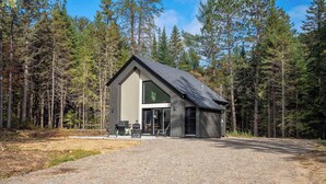 A modern cabin with a large glass window, a gravel driveway, and a forested backdrop.