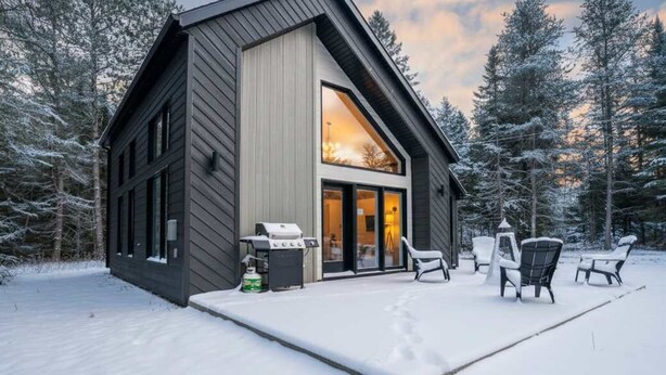 A modern cabin with a large glass window, a patio with outdoor furniture, and a snow-covered landscape.