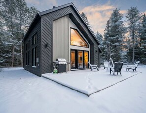 Una cabaña moderna con un amplio ventanal, un patio con mobiliario exterior y un paisaje cubierto de nieve.