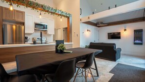 A modern kitchen with stainless steel appliances, a dining area with a dark table and chairs, and a living room with a black sofa and a flat-screen TV.