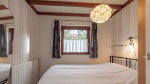 A bedroom with a large bed, a window with curtains, a radiator, and a hanging light fixture. - Orchis4 Bungalow Near Dunes in Julianadorp (Julianadorp)