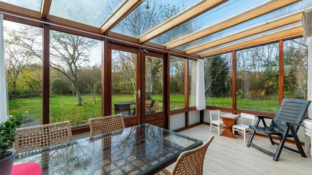A conservatory with a glass table and wicker chairs, surrounded by wooden frames and large windows. - Orchis4 Bungalow Near Dunes in Julianadorp (Julianadorp)
