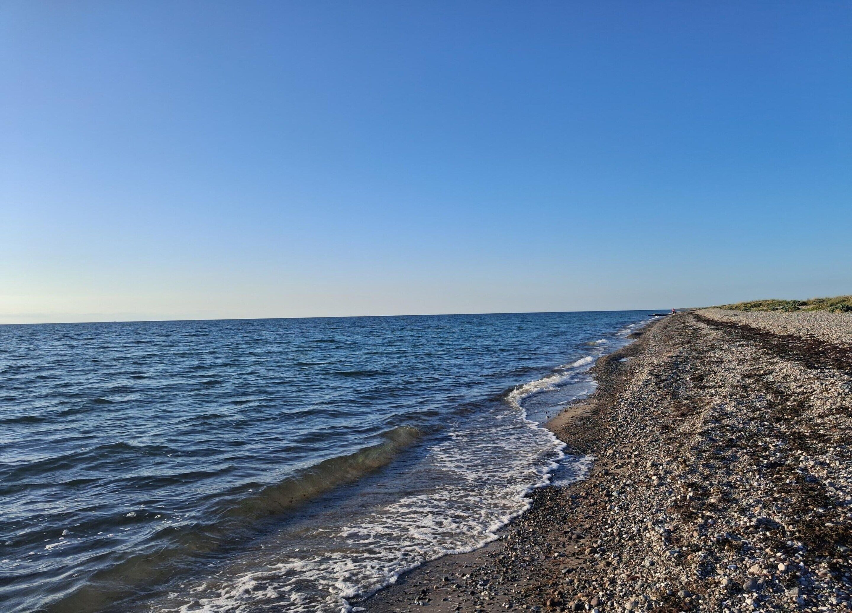 Ein Kiesstrand mit Wellen, die an die Küste schlagen, unter blauem Himmel.