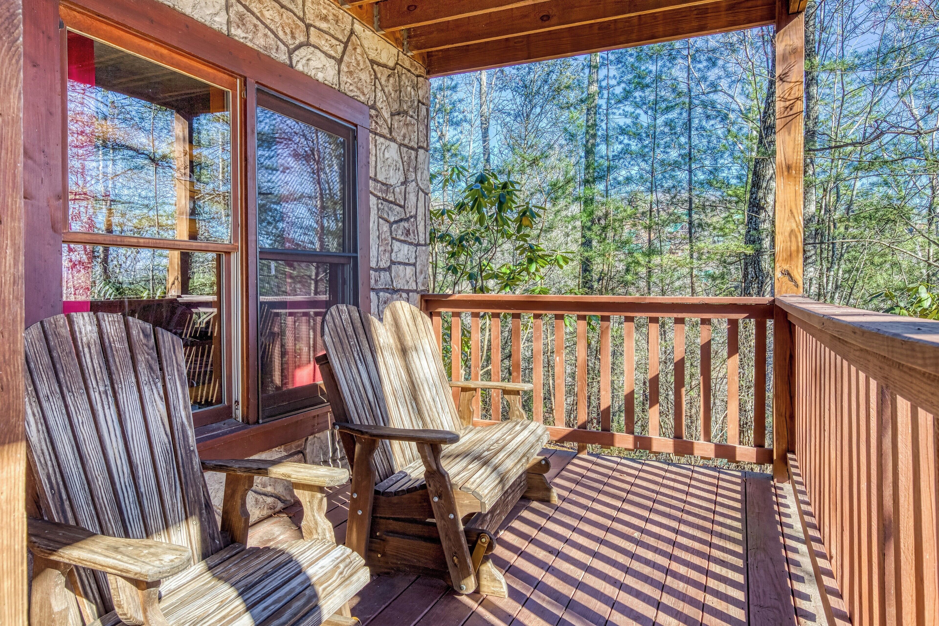 A wooden deck with two Adirondack chairs, a stone wall, and a view of a forest.