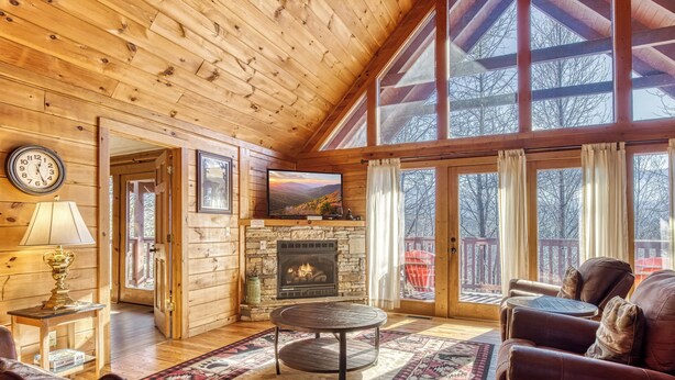 A cozy cabin interior with a wooden ceiling, a fireplace, a television, leather furniture, and large windows. - ABOVE THE CLOUDS! 4 Bedroom in the Smokies! (Gatlinburg)