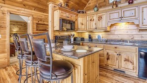 A wooden cabin interior with a kitchen featuring granite countertops, wooden cabinets, and a central island with bar stools. - ABOVE THE CLOUDS! 4 Bedroom in the Smokies! (Gatlinburg)