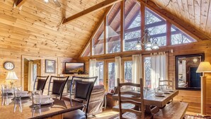A wooden cabin interior with a dining area, kitchen, and large windows. - ABOVE THE CLOUDS! 4 Bedroom in the Smokies! (Gatlinburg)