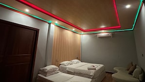 A modern bedroom with two beds, a wooden wardrobe, a seating area, and a ceiling with red and green LED lighting. - NikainnHimandhoo (Himandhoo)