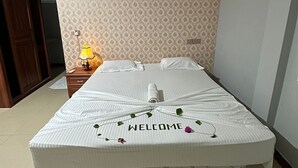 A neatly made bed with a 'WELCOME' sign and decorative flowers, a bedside lamp, and an air conditioning unit on the wall. - NikainnHimandhoo (Himandhoo)