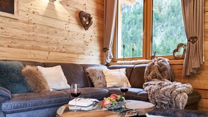 A cozy cabin interior with a leather sofa, a wooden coffee table with fruits and wine glasses, and a large window with curtains. - Chalet in Leutasch Near Ski Trails (Leutasch)