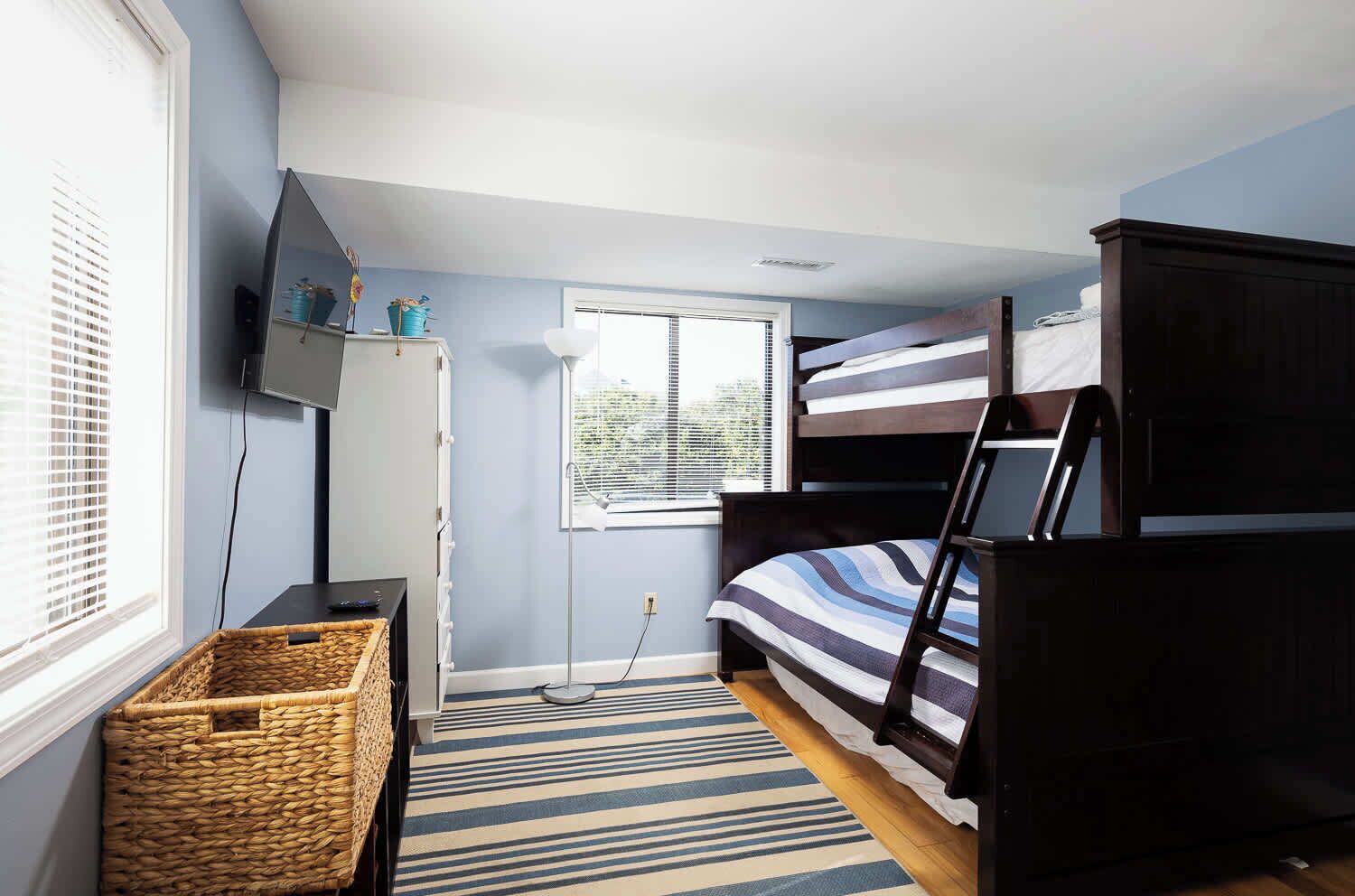 Een slaapkamer met een stapelbed, een bureau, een lamp, een raam en een rieten mand.