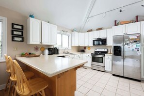 A modern kitchen with white cabinets, a stainless steel refrigerator, a built-in oven, and a large island with bar stools. - Semi-Oceanfront w/ Heated Pool & Cabana (Corolla)