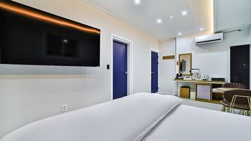 A modern hotel room with a large bed, a flat-screen TV mounted on the wall, a desk with a chair, and a small kitchenette.
