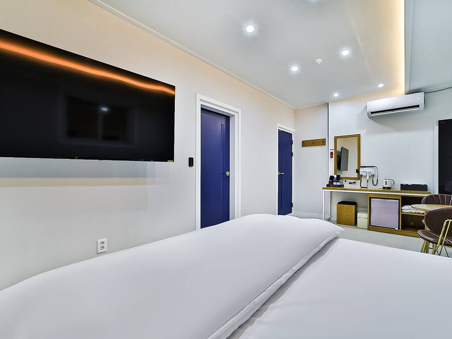 A modern hotel room with a large bed, a flat-screen TV mounted on the wall, a desk with a chair, and a small kitchenette.