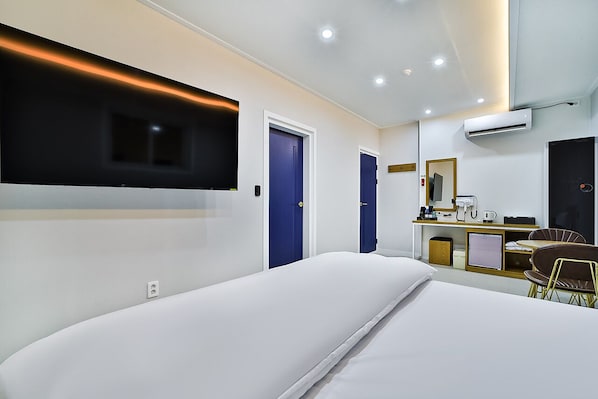 A modern hotel room with a large bed, a flat-screen TV mounted on the wall, a desk with a chair, and a small kitchenette.