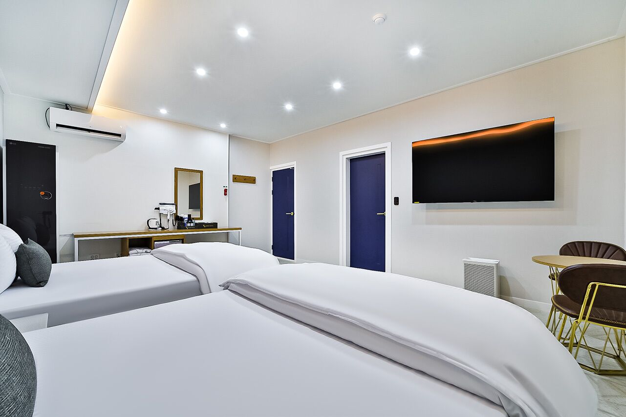 Habitación de hotel con dos camas, un escritorio con silla, televisor montado en la pared y una pequeña mesa de comedor con sillas.