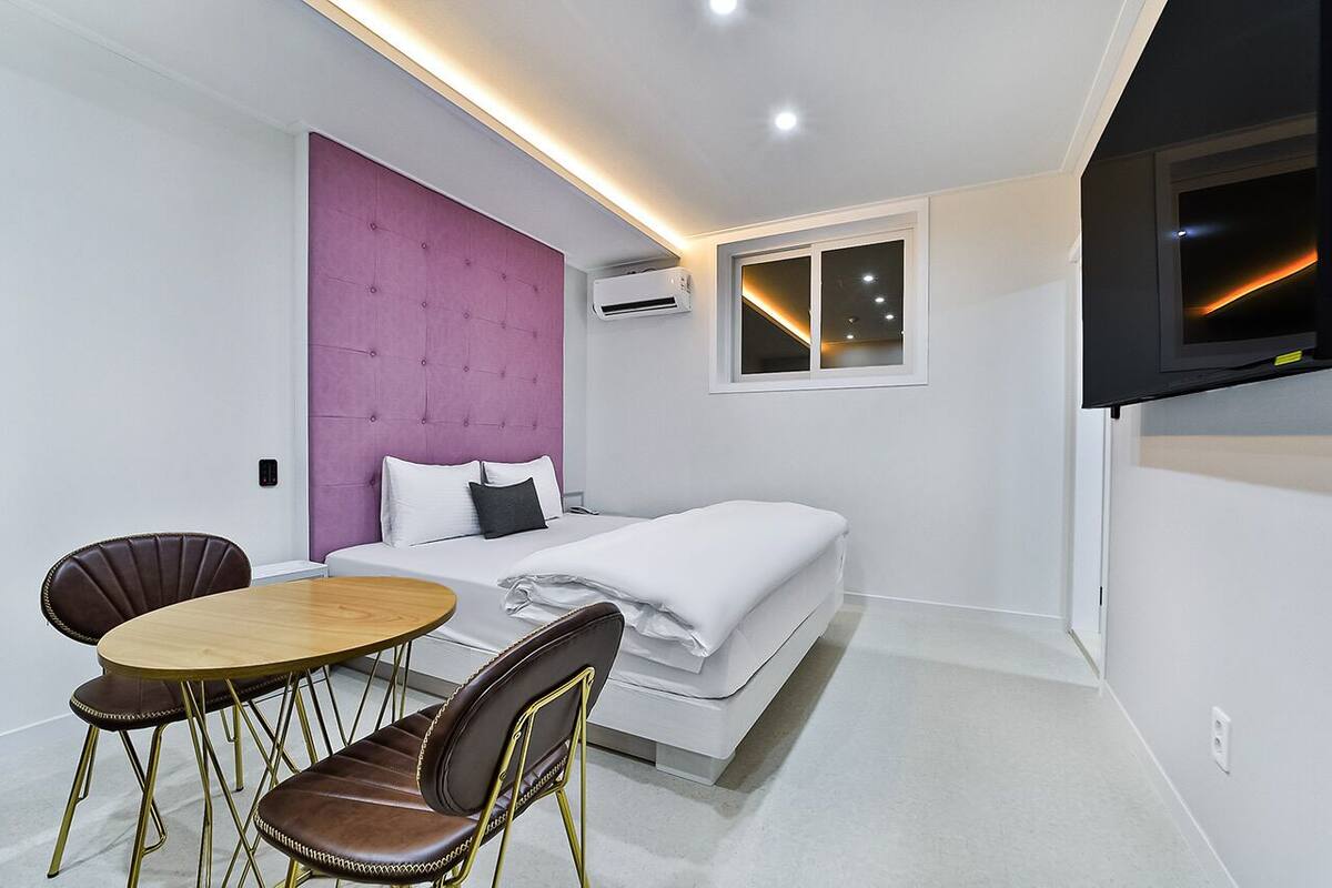 A modern hotel room with a bed, a small table, two chairs, a TV, and an air conditioning unit.