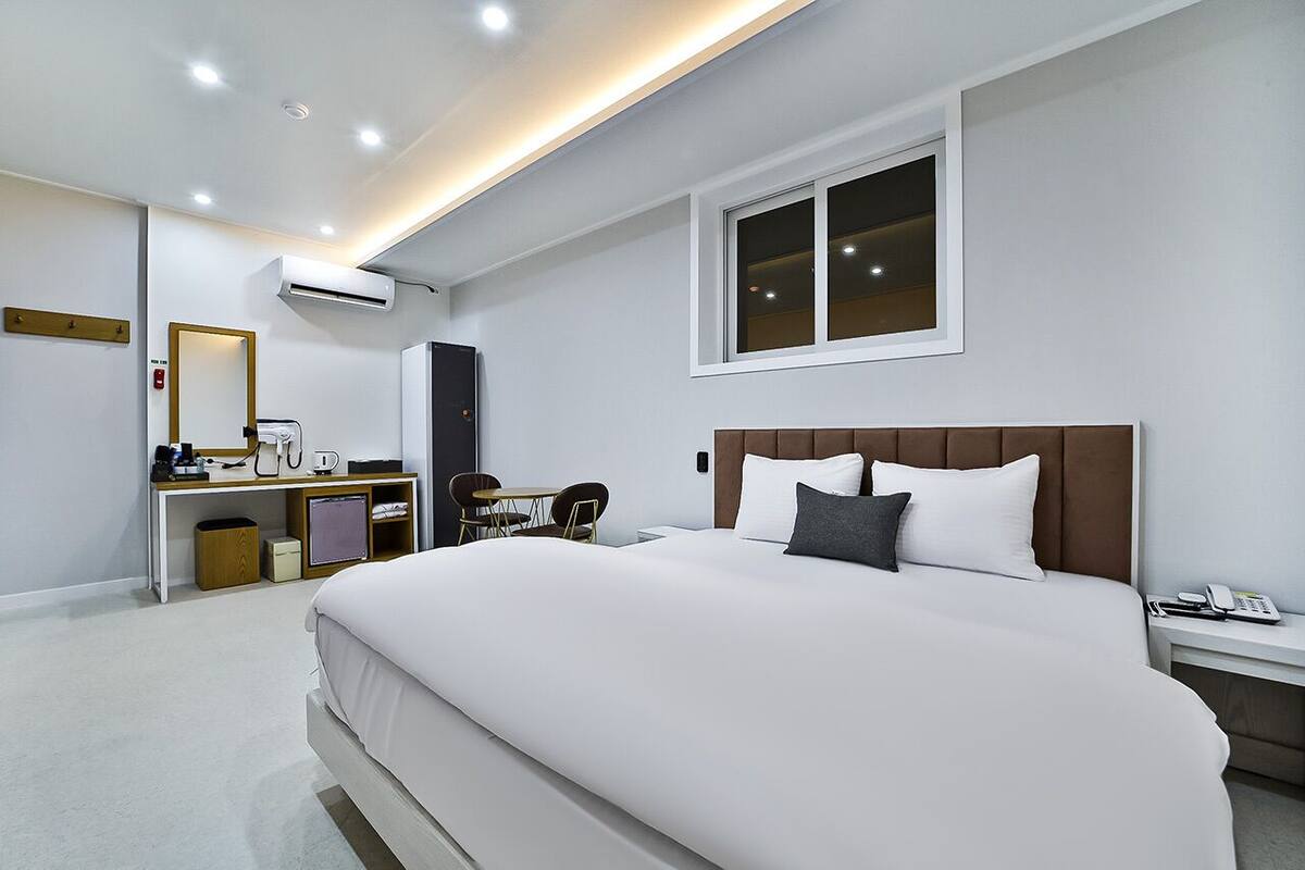 A modern hotel room with a large bed, a desk with a chair, a small table, and a window.