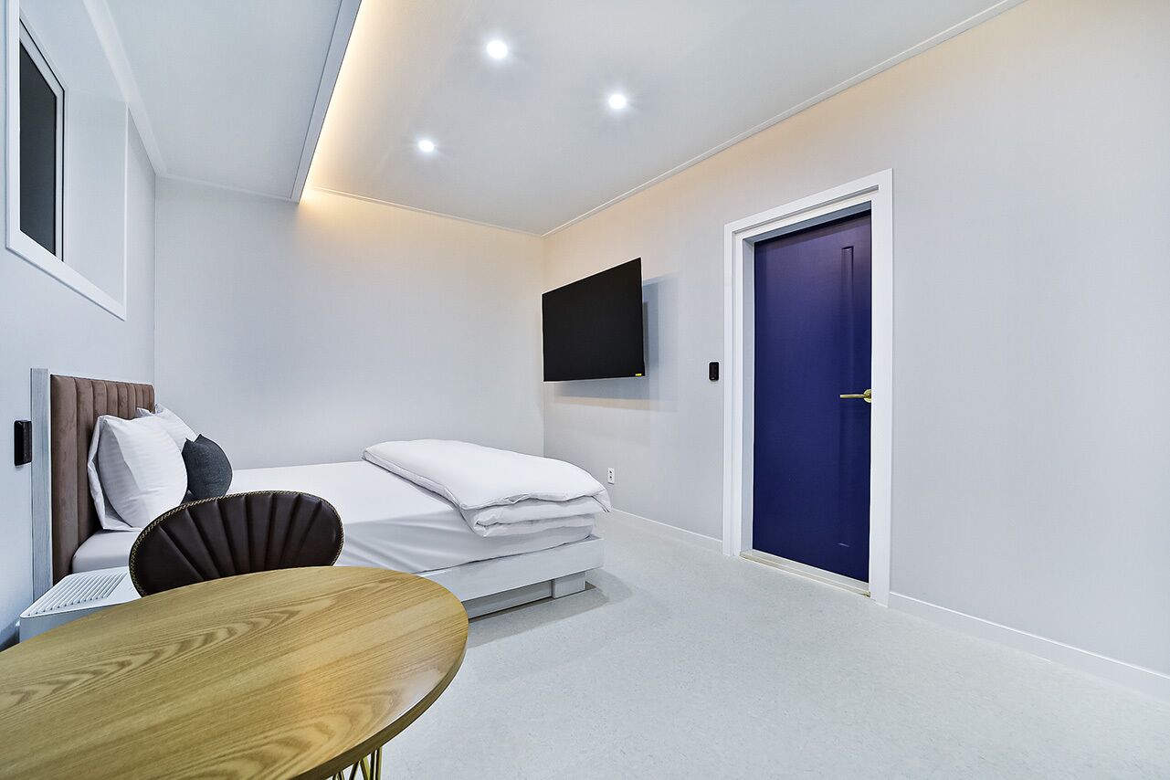 Una habitación de hotel moderna y funcional con una cama, un televisor de pantalla plana montado en la pared, una mesa de madera y una puerta azul.