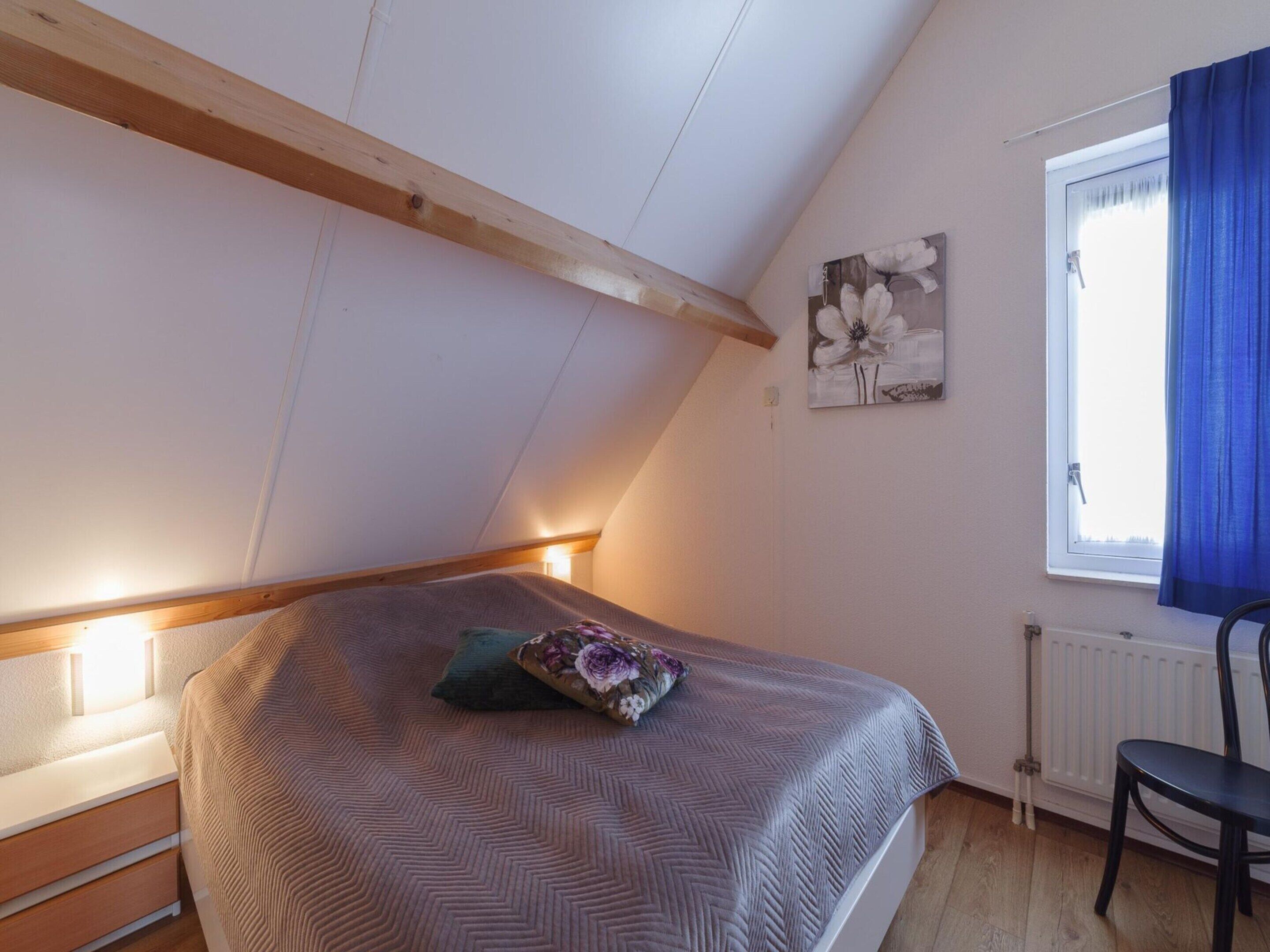 Ett sovrum med ett sluttande tak, en säng med ett mönstrat överkast, en kudde, en väggmonterad lampa, ett element, ett fönster med blå gardiner och en stol.