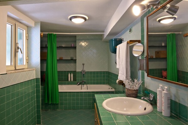 A bathroom with a green tiled floor, a white sink, a bathtub, a green shower curtain, a mirror, and a window with a frosted glass door.