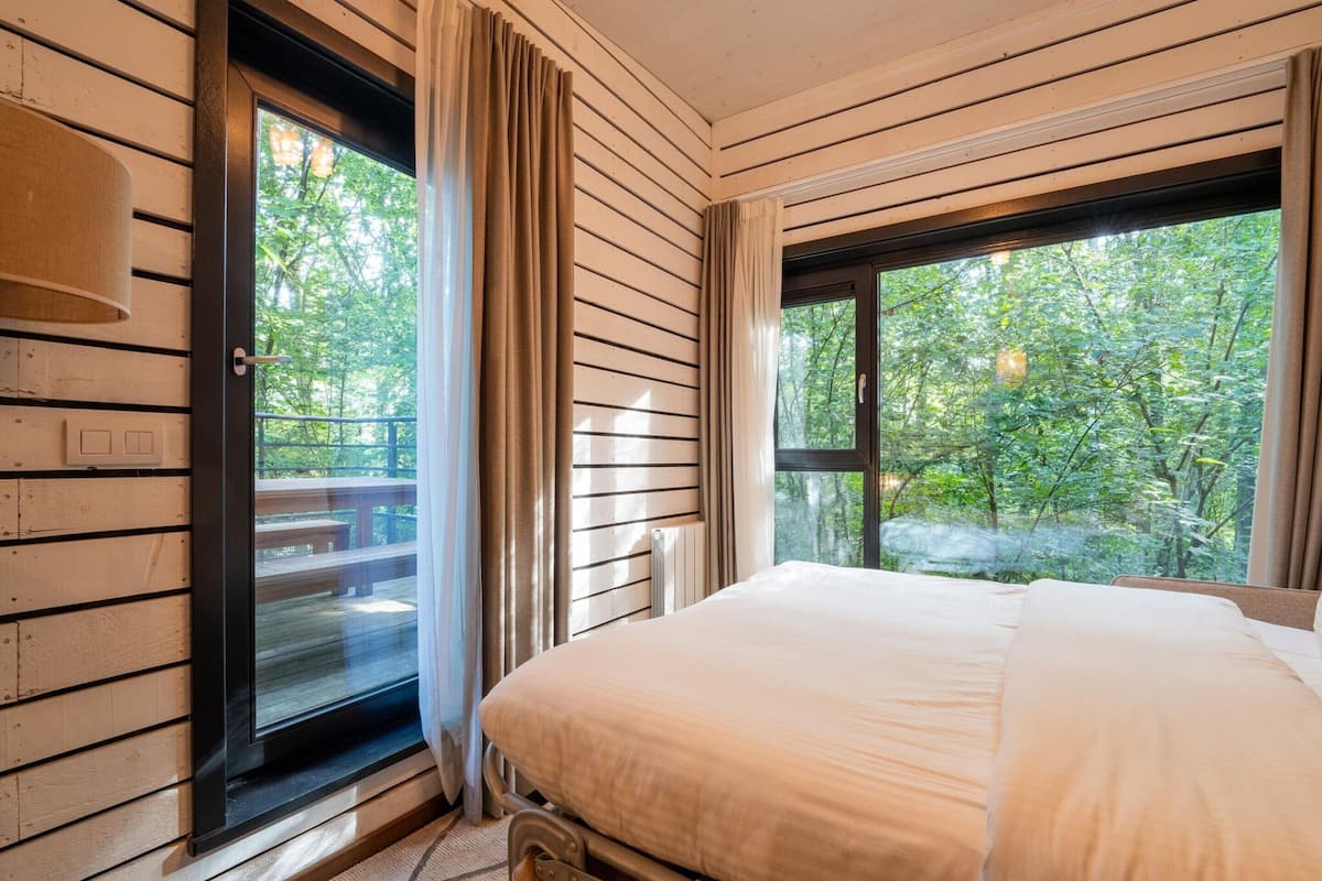 A bedroom with a large bed, wooden walls, a window with curtains, and a view of trees.