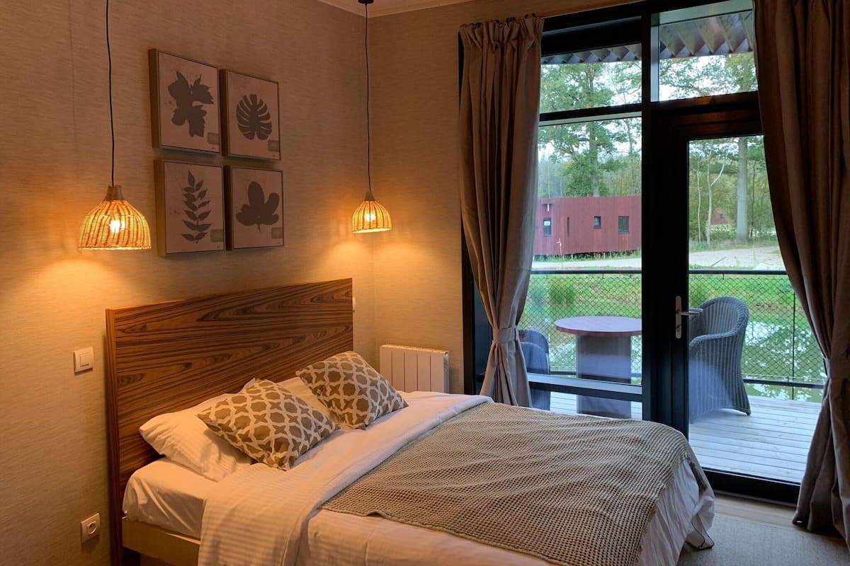 A bedroom with a wooden headboard, a bed with patterned pillows, two pendant lights, and a view of a deck and greenery outside.