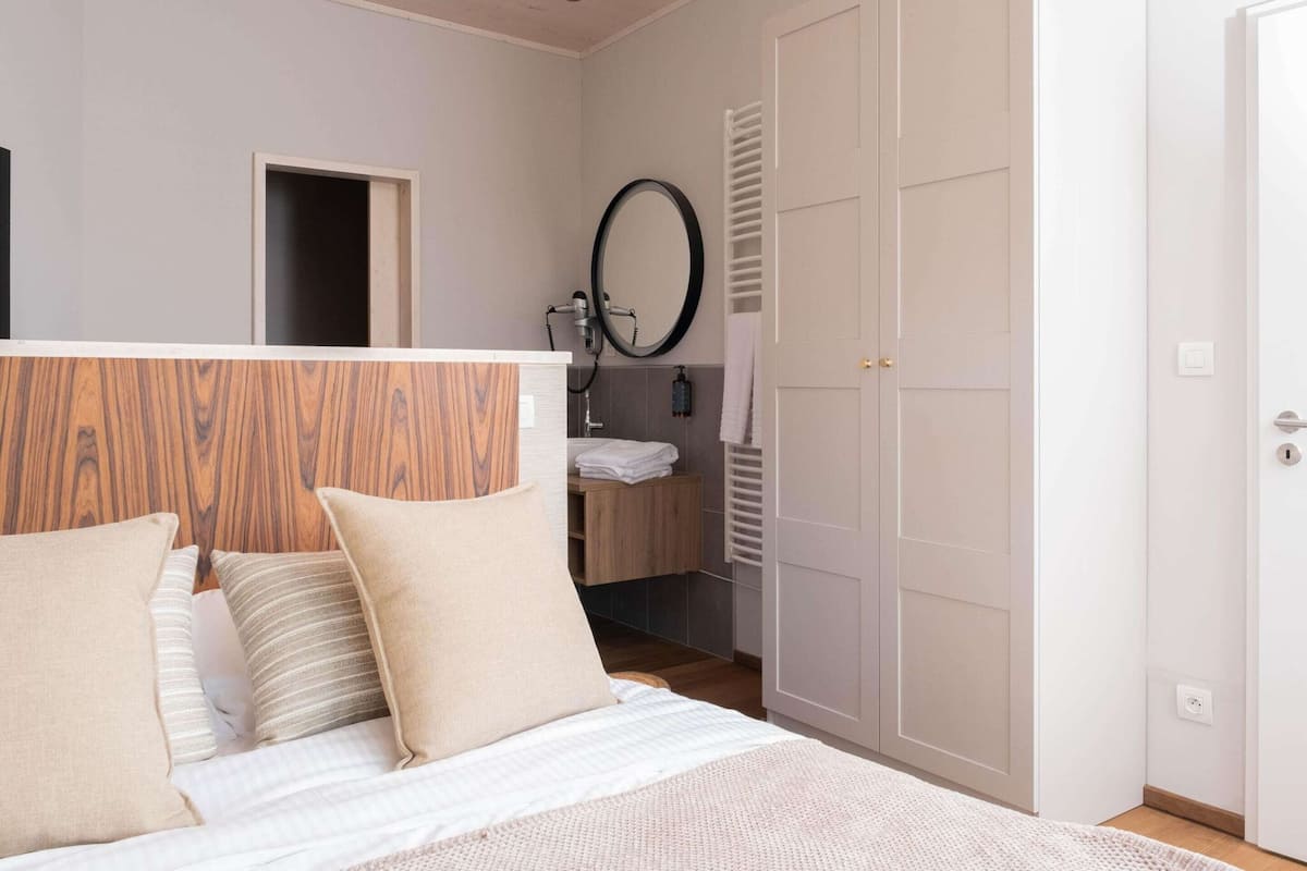 A bedroom with a wooden headboard, a bed with white bedding, a bedside table, a mirror, a wardrobe, and a bathroom with a towel rack.