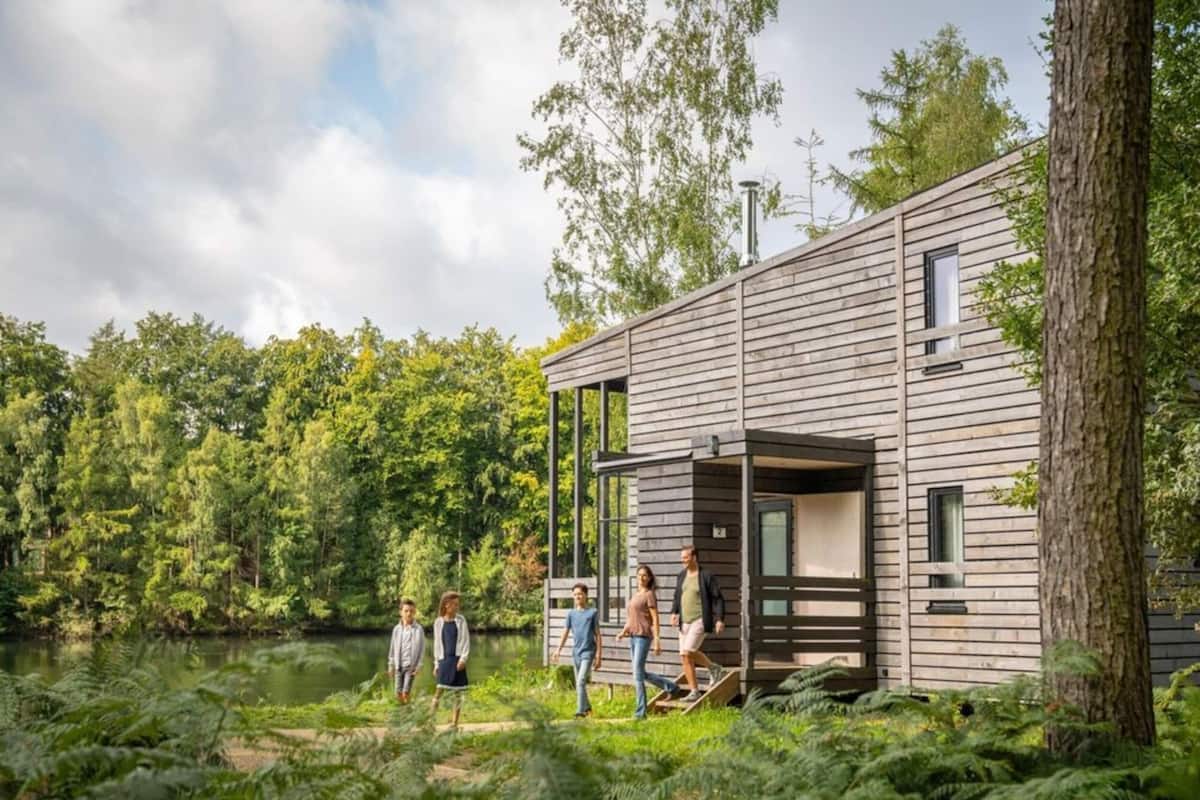 A modern wooden cabin with a deck, surrounded by dense forest and a lake.