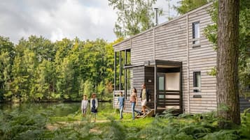 A modern wooden cabin with a deck, surrounded by dense forest and a lake.