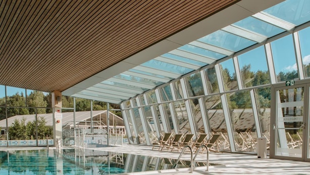 An indoor swimming pool with clear water, wooden ceiling, and glass walls offering views of the outdoors. - Lakeside Luxury Mansion (Antoing)