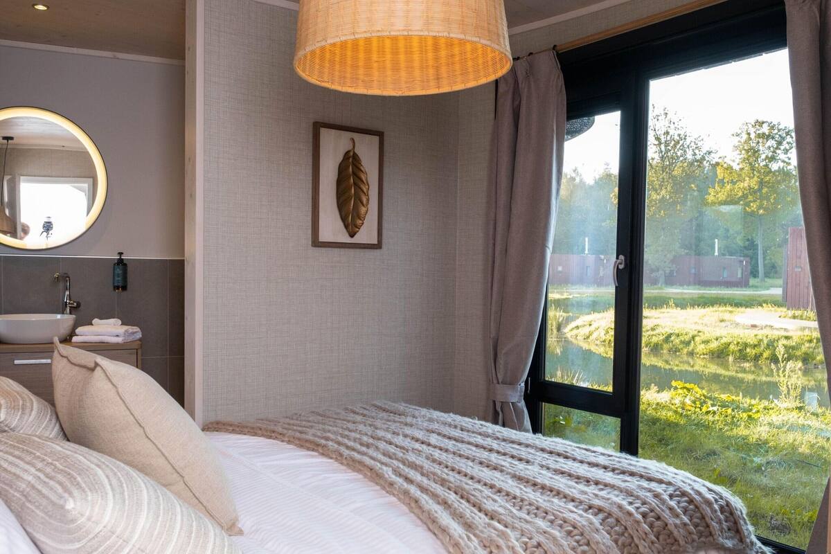 A bedroom with a large bed, a round mirror, a framed leaf artwork, and a window with a view of greenery and a building.
