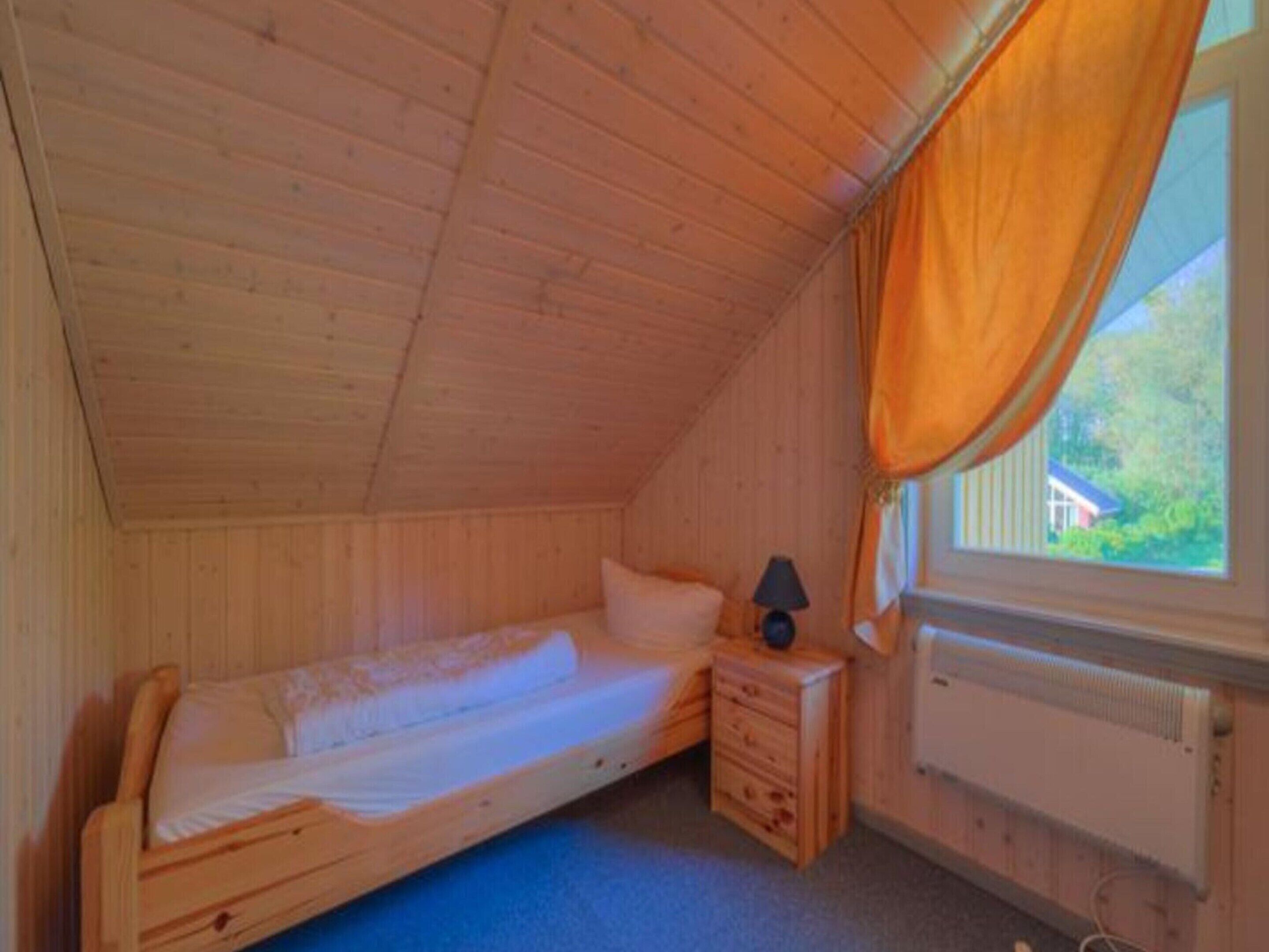 A small, single-bed room with wooden walls and a sloped ceiling. There is a window with an orange curtain, a bedside table with a lamp, and a radiator.