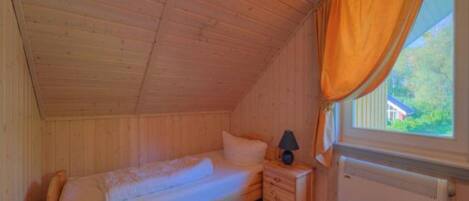 A small, single-bed room with wooden walls and a sloped ceiling. There is a window with an orange curtain, a bedside table with a lamp, and a radiator.