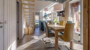 A modern interior with a wooden staircase, a dining area with a table and chairs, and a large window with curtains. - Ferienhaus im Skandinavischen Stil (Rechlin)