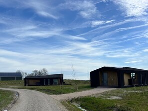 Un bâtiment moderne avec une allée de gravier menant à une porte en bois, entouré de champs ouverts et surplombé d’un ciel dégagé.