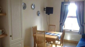 Een kamer met een eettafel, stoelen, een aan de muur bevestigde televisie, een raam met gordijnen en een blauwe bank.