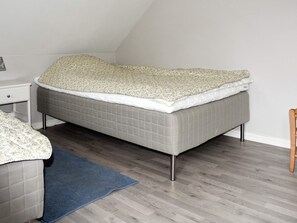 Une chambre avec un lit simple, un couvre-lit gris, un tapis bleu et une chaise en bois.