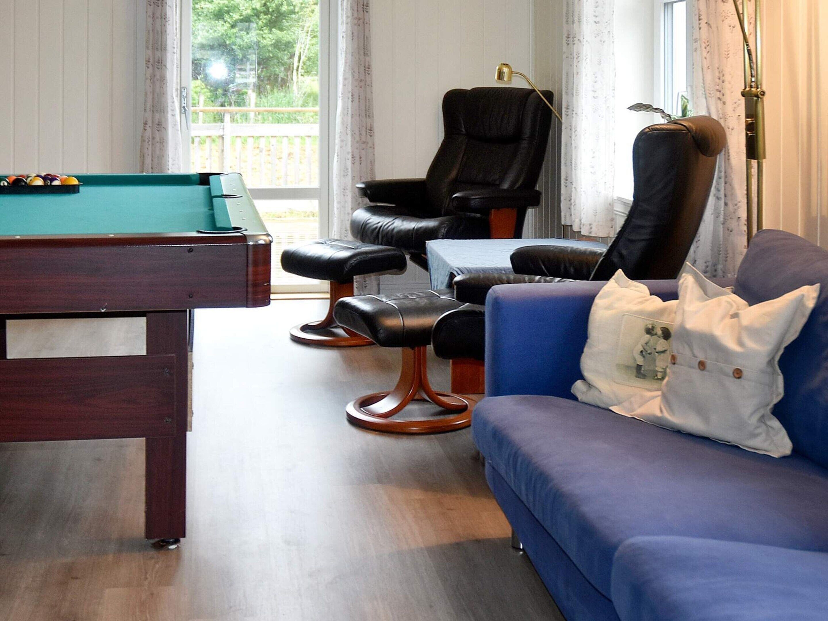 A room with a pool table, two leather chairs, a blue sofa, and a window with a view of greenery.
