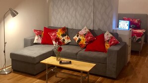 Ein modernes Wohnzimmer mit einem grauen Sofa, roten und floralen Kissen, einem Holztisch, einer Stehlampe und einem Schild mit der Aufschrift 'Welcome to Graz'.