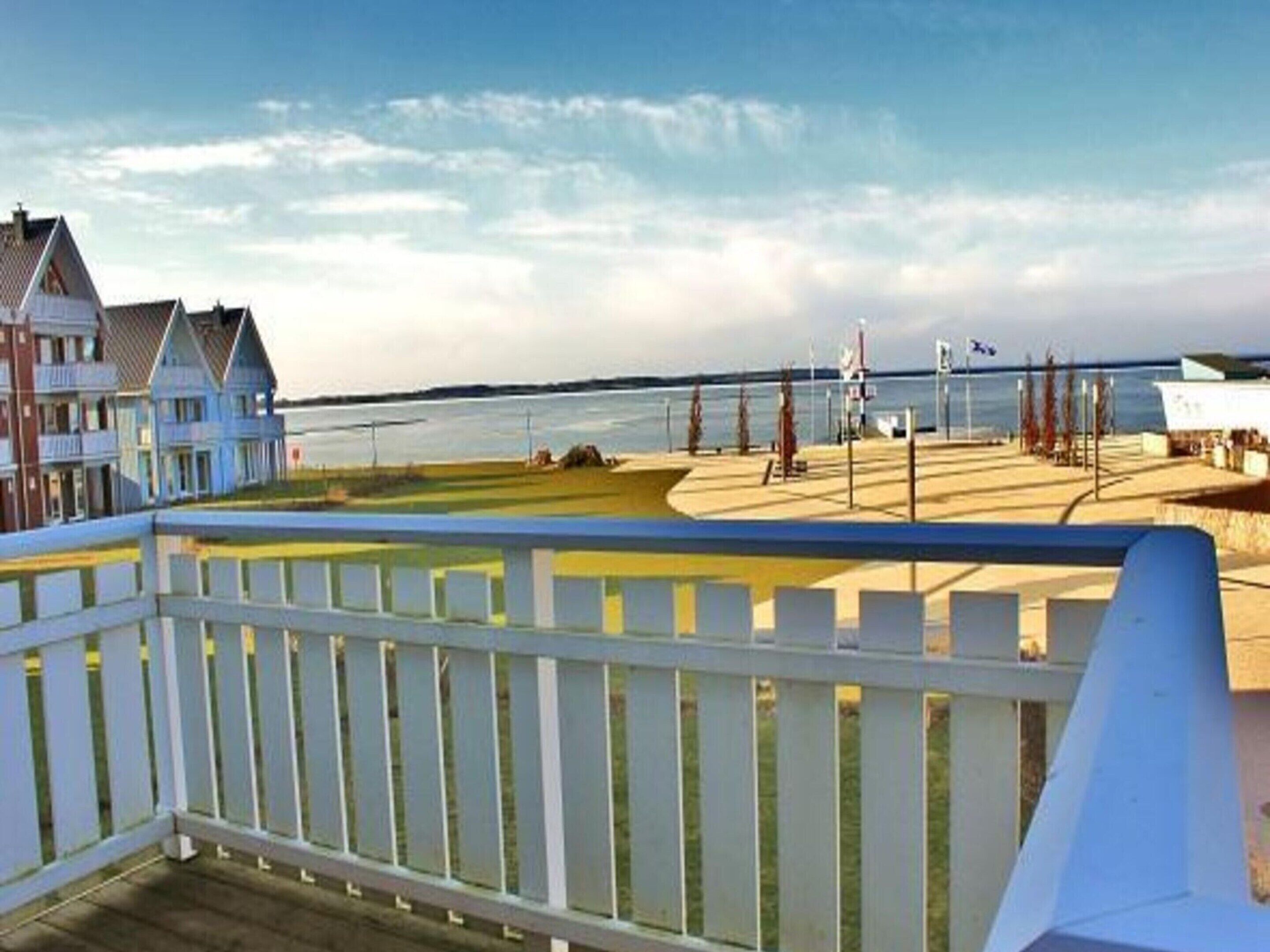 A coastal view from a balcony with a white railing, overlooking a beach with a sandy area, a few trees, and a body of water.