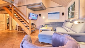 Ein modernes Wohnzimmer mit einer Holztreppe, einem großen grauen Sofa, einem kleinen Tisch mit Pflanze und einem flachbildfernseher an der Wand.