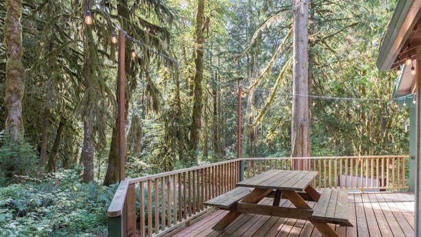 A wooden deck with a picnic table surrounded by a wooden railing, set in a forested area with tall trees and hanging lights. - Summer special, stay before 9/8/26 get third night free. (Welches)