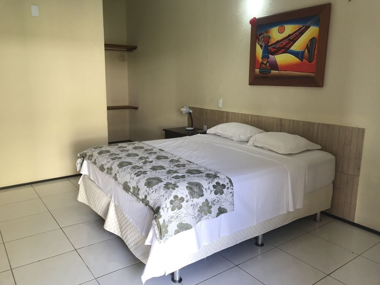 A neatly made bed with a floral bedspread, a bedside table with a lamp, and a painting on the wall.