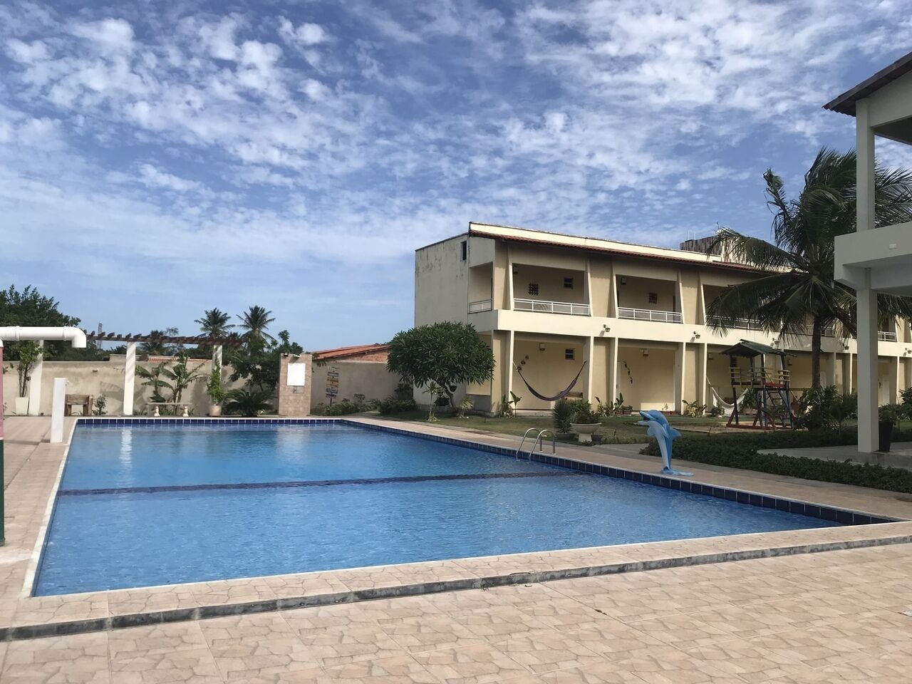 A swimming pool with a dolphin sculpture, surrounded by a building with balconies and palm trees.