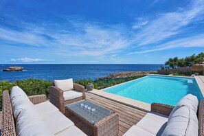 A poolside area with wicker furniture, a clear view of the ocean, and a wooden deck. - Lago Spectacular Villa on the Seafront (Cala D'or)