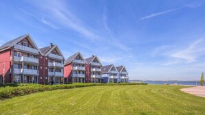 A row of red and white two-story buildings with balconies, a well-maintained lawn, and a clear view of the sea in the background. - Apartment in Rechlin by Muritz National Park (Rechlin)